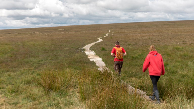Two people in red coats walking down a narrow, man-made, pathway on top of moorland.